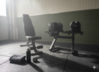 Moody home gym corner with the WOLV bench, racked adjustable dumbbells, towel and bottle on rubber tiles